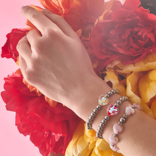 Crimson Swirl Glowing Heart Essence Bracelets with silver beads and glowing heart charms worn on wrist against red and yellow flowers by NOGU